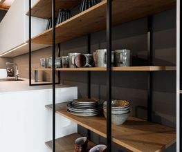 Moderne, hochwertige Designküche mit weißer Kücheninsel, dunklen Glasfronten und offenem Regal aus massivem Holz. Handgefertigtes Keramikgeschirr in natürlichen Grau- und Brauntönen setzt warme Akzente und unterstreicht den minimalistischen, modernen Wohnstil.