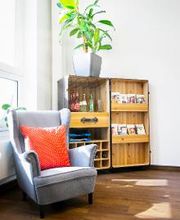 gemütlicher Ohrensessel im Coachingzimmer. Dahinter ein großes Fenster wodurch viel Tageslicht scheint sowie ein Regal mit Broschüren und Getränken. Darauf steht eine große Zimmerpflanze.