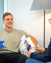 Teilnehmergespräch, Mann und Frau sitzen sich gegenüber. Mann hält blaue Tasse, Frau einen Flyer. Hinter ihnen steht eine Stehlampe.