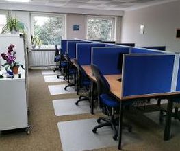 Ein Büroraum mit mehreren Arbeitsplätzen, die durch blaue Trennwände abgetrennt sind. Jeder Platz hat einen Bürostuhl und eine Schreibtischmatte. Links steht ein Regal mit einer Orchidee. Große Fenster lassen Tageslicht herein, draußen sind Bäume zu sehen