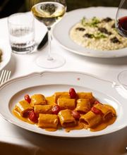 Elegant plate of stuffed pasta coated in a rich orange sauce and garnished with cherry tomatoes, set on a white tablecloth with wine glasses and a truffle risotto visible in the background.