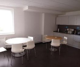 Ein Pausenraum mit zwei runden Tischen und weißen Stühlen. Rechts ist eine Küchenzeile mit weißen Schränken, Kaffeemaschinen und einer Mikrowelle. Ein großes Fenster lässt Tageslicht herein, und der Boden ist dunkelgrau.