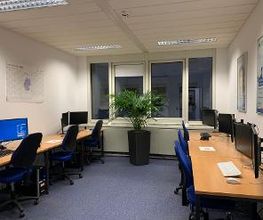 Schulungsraum mit mehreren Arbeitsbereichen, die jeweils mit 2 PC-Bildschirmen, Tastatur, Maus und Headset ausgestattet sind. Große Zimmerpflanze vor der Fensterfront. Bilder und Jahreskalender an der Wand.