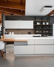 Großzügige, moderne Designküche mit weißen, grifflosen Küchenfronten, einer zentralen Kochinsel und warmen Holzelementen. Die offene Regalwand mit dunklem Metallrahmen, die sichtbare Holzdecke und die hochwertige Ausstattung verbinden minimalistisches Küchendesign mit natürlicher Wohnlichkeit.