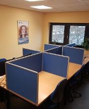 Schulungsraum mit mehreren Arbeitsbereichen, die durch blaue Trennwände voneinander abgegrenzt sind und mit PC-Bildschirmen, Tastatur, Maus und Headset ausgestattet sind. Fensterreihe und Bilder an der Wand.