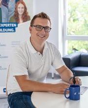 junger Mann mit Brille sitzt an einem Tisch und füllt ein Formular aus. Vor ihm steht eine Tasse.