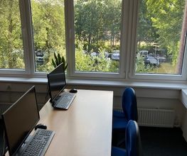 2 Arbeitsplätze vor einer Fensterfront mit Blick ins Grüne. Auf den Tischen befinden sich 2 Computerbildschirme mit dazugehöriger Tastatur und Maus.