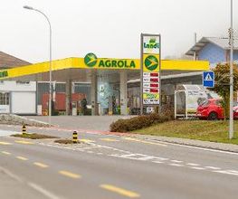 AGROLA Tankstelle in Triengen mit TopShop; im Vordergrund Strasse und Fussgängerstreifen. Grosse Überdachung und Totem mit Landi Logo, AGROLA Diskus und digitaler Preisanzeige und TopShop Logo 