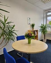 Ein heller Raum mit einem runden Holztisch, darauf ein Blumenstrauß in einer grünen Vase. Um den Tisch stehen blaue Stühle. Im Hintergrund stehen Grünpflanzen, ein kleines Regal und ein Fenster, das Tageslicht hereinlässt. Die Wände sind weiß.