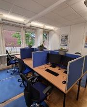 Ein Bild von den Arbeitsplätzen, welche mit blauen Trennwänden voneinander getrennt sind, mit 2 Bildschirmen, Tastatur, Maus sowie Headset ausgestattet sind. Die dazugehörigen Bürostühle stehen auf blauen Schutzmatten. 