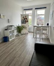 ein heller Aufenthaltsraum mit 2 großen Fenstern. Rechts steht ein weißer Tisch mit Bar-Hockern. Links steht ein Regal, auf dem ein Drucker steht. Daneben steht eine Zimmerpflanze und ein Roll Up der WBS GRUPPE. Im Vordergrund ist eine schwarze Couch