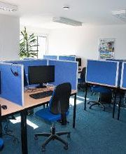 Dieses Bild zeigt einen Schulungsraum. Zu sehen sind Schreibtische, welche jeweils mit zwei Bildschirmen, Tastatur, Maus und Headset ausgestattet sind.