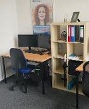 2 Arbeitsplätze mit jeweils 2 Bildschirmen, einem Bürostuhl, einer Tastatur und Maus. Darüber hängen 2 Plakate, zwischen den Tischen steht ein Regal mit Ordnern.