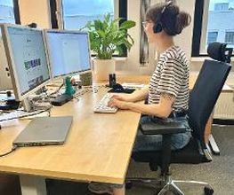 Arbeitsplatz mit 2 Bildschirmen, Tastatur und Maus. Eine Frau sitzt auf einem Bürostuhl am Tisch und arbeitet. Im Hintergrund steht eine Zimmerpflanze.