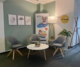 Eingangsbereich mit grüner Wand an der 3 Bilder mit Pflanzen hängen. Im Raum steht ein Beistelltisch mit 3 grauen Sesseln. Dahinter steht ein Banner und eine Stehlampe. Rechts ein Kleiderständer und eine Pflanze. 