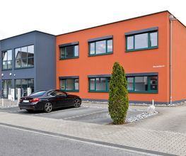 Außenansicht eines orange-grauen Gebäudes, im Vordergrund Parkplatz mit einem schwarzen PKW und ein Baum.