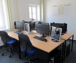 Schulungsraum mit 4 Arbeitsplätzen mit jeweils 2 Bildschirmen, einem Bürostuhl, Tastatur, Maus und Headset. An der Wand hängt ein Wandkalender.