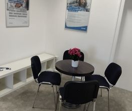 Sitzecke. Ein schwarzer Tisch mit Blumen und 4 Stühlen. Im Hintergrund hängen Poster an der Wand.