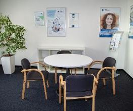 Beratungszimmer. Ein runder Tisch mit vier Stühlen. Im Hintergrund steht eine grüne Zimmerpflanzen und es hängen verschiedene Plakate an der Wand.