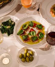 A table spread featuring a bowl of tuna crudo garnished with basil leaves, accompanied by green olives, sautéed greens, red wine, and a orange cocktail.