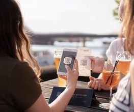 Zwei Frauen sitzen in einem Außenbereich und scannen mit ihren Smartphones einen QR-Code, während sie Getränke genießen. Das Bild zeigt die nahtlose Integration digitaler Technologien von gastronovi wie QR-Code-Bestellsysteme zur Selbstbedienung in der Gastronomie.