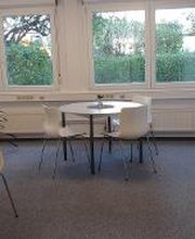 Ein heller Pausenraum mit runden Tischen und weißen Stühlen. Auf den Tischen stehen kleine Dekorationen. Rechts ist ein Regal mit bunten Ordnern, links Pflanzen. Große Fenster lassen Licht hinein und bieten Blick auf Büsche und Bäume. Der Boden ist grau.