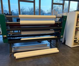 mediaService Bremen | Full-Stack Werbeagentur - Eine große professionelle Rollen-Laminiermaschine in einer Werkstatt für Werbetechnik, bestückt mit mehreren breiten Papier- und Folienrollen zur Weiterverarbeitung.