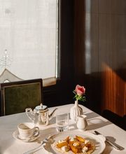 Morning sunlight illuminates a table set with a silver teapot, teacup, pink flower, and a thin crepe topped with caramelized fruit and whipped cream.