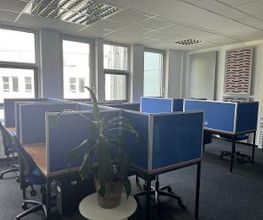 Ein Büro mit mehreren Arbeitsplätzen, die durch blaue Trennwände unterteilt sind. Jeder Arbeitsplatz hat einen Bürostuhl. Vorne steht eine grüne Pflanze auf einem kleinen Tisch. Große Fenster lassen viel Tageslicht hinein.