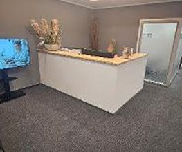 Empfangsbereicht mit modernem Thresen, 2 Sesseln, einem großen Fernseher auf dem ein standbild mit Informationen zu sehen ist. Mehrere Topfpflanzen. Blick in den Flur.