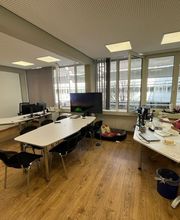 Büroraum mit 3 Schreibtischen vor einer großen Fensterfront. An einem Schreibtisch sitzt eine Mitarbeiterin vor ihrem PC-Bildschirm.