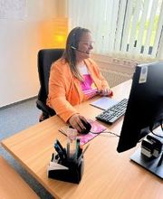 Eine Frau mit orangener Jacke sitzt an einem PC. Sie hat ein Headset auf dem Kopf.