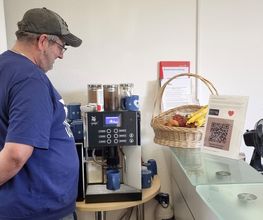 Person steht an Kaffeemaschine, daneben eine Theke mit frischem Obst im Korb und dem Aufsteller mit QR-Code.
