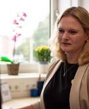 Eine blonde Frau in beigem Blazer sitzt an einem Schreibtisch und schaut auf einen Monitor. Auf der Fensterbank hinter ihr stehen eine Orchidee und gelbe Blumen. Durch das Fenster ist unscharf eine grüne Umgebung zu sehen. Der Raum ist hell und ruhig.