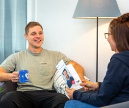 Teilnehmergespräch, Mann und Frau sitzen sich gegenüber. Mann hält blaue Tasse, Frau einen Flyer. Hinter ihnen steht eine Stehlampe.