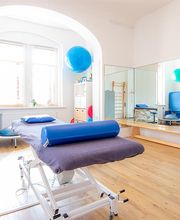 Ein großzügiger und lichtdurchfluteter Physiotherapie-Raum mit einer zentral stehenden höhenverstellbaren Therapieliege mit dunkelgrauem Bezug und blauen Polsterrollen. Der Boden ist aus hellem Holz, und die Wände sind neutral weiß gehalten. Links steht ein medizinisches Gerät auf einem weißen Rollwagen, vermutlich für Elektro- oder Ultraschalltherapie. An der Wand befindet sich ein großer Spiegel, der fast die gesamte Breite einnimmt, um Bewegungsabläufe beobachten zu können. Rechts sind blaue Gymnastikbälle an der Wand befestigt. Ein Regal mit Therapie- und Fitnessutensilien wie Balancekissen, Hanteln und Dehnungsbändern steht ebenfalls im Raum. Große Fenster mit weißen Jalousien lassen viel Tageslicht herein.