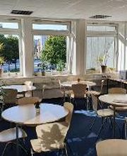 Ein heller Pausenraum mit runden Tischen, Holzstühlen und kleinen Pflanzen darauf. Große Fenster lassen Tageslicht herein und bieten Blick auf Bäume und die Straße. Grünpflanzen stehen im Raum, rechts befinden sich Schränke und Geräte. Der Boden ist blau.