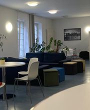 Aufenthaltsraum mit blauen Sofas, weißen Tischen und Stühlen, einem Bücherregal, großen Fenstern und einigen Zimmerpflanzen.