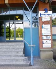 Außenansicht des Eingangsbereich vom Gebäude. Glas Eingangstür mit blauen Rahmen. Rechts an der Seite hängt eine Schildertafel der ansäßigen Unternehmen.