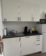 eine weiße Einbauküche mit 6 Schränken und einer Spüle links. Rechts stehen Wasserkocher und eine Kaffeemaschine. Neben der Küche steht ein Kühlschrank, auf dem eine Mikrowelle steht. 