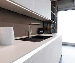 Diese elegante LEICHT Designküche überzeugt mit einer puristischen Gestaltung, hochwertigen Materialien und einer durchdachten Lichtinszenierung. Die weiße, grifflose Arbeitszeile wird durch eine schwarze Einbauspüle und eine edle Edelstahlarmatur stilvoll ergänzt. Indirekte LED-Beleuchtung unter den Oberschränken sorgt für eine angenehme Arbeitsatmosphäre und setzt die klaren Linien der Küche gekonnt in Szene. Offene Designregale mit schwarzen Metallrahmen verleihen der modernen Wohnküche zusätzliche Leichtigkeit und bieten Platz für stilvolle Dekoration.