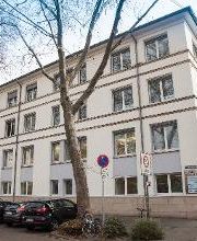 Seitliche Außenansicht auf das Gebäude von der gegenüberliegenden Straßenseite. Hier wird die Ansicht von der Hauptstraße auf das seitliche Gebäude präsentiert. Bäume und Parkmöglichkeiten.