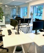 Hell und offen gestalteter Schulungsraum mit weißen, modernen Schreibtischen und schwarzen, modernen Schreibtischstühlen. Monitore auf den Tischen. Helle Glasfront sorgt für viel Licht. Heller Holzfußboden. Grünpflanze.