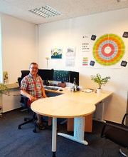 Ein Mitarbeiter sitzt in seinem hellen Büro an seinem Schreibtisch und lächelt sympathisch in die Kamera.