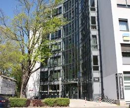 Außenansicht vom Gebäude. Helle Fassade mit vielen Fenstern. Im Vordergrund steht ein großer Baum.