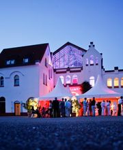 Die Palazzo Halle in Karlsruhe – Ihre perfekte Eventlocation für unvergessliche Veranstaltungen wie Firmenfeiern, Produktpräsentationen, Tagungen, Hochzeiten und mehr.
Die ehemalige Maschinenfabrik am Westbahnhof in Karlsruhe bildet ein außergewöhnliches Ambiente für exklusive Veranstaltungen mit dem gewissen Etwas. Auf 1.000 Quadratmetern ermöglicht die flexible Raumeinteilung den passenden Rahmen für jede Art von Event – von privaten Feiern wie Hochzeiten oder Geburtstage bis hin zu geschäftlichen Events wie Tagungen, Konferenzen oder Produktpräsentationen. In den Abendstunden taucht die ausgeklügelte Beleuchtungstechnik die hohen Decken in unterschiedliche Lichtstimmungen und macht die Palazzo Halle zu einer einmaligen Eventkulisse. Die Halle kann über die Eventagentur Projekt Eins gemietet werden, die das Lichtkonzept, die Dekoration, das Catering und auf Wunsch auch ein Rahmenprogramm detailliert auf das jeweilige Thema der Veranstaltung abstimmt.