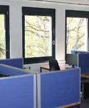 Lernplätze die durch blaue Trennwände voneinander getrennt sind. Jeder Lernplatz besteht aus 2 PC-Bildschirmen, Tastatur und Maus sowie Headset. Fensterfront mit Blick ins Grüne.