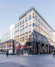 Außenansicht des Gebäudes. Helle Fassade und viele Fenster. Belebte Umgebung.