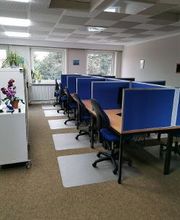 Ein Büroraum mit mehreren Arbeitsplätzen, die durch blaue Trennwände abgetrennt sind. Jeder Platz hat einen Bürostuhl und eine Schreibtischmatte. Links steht ein Regal mit einer Orchidee. Große Fenster lassen Tageslicht herein, draußen sind Bäume zu sehen