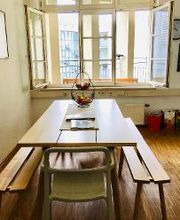 Die gemütliche Küche, die Mitarbeiter:innen und Teilnehmer:innen für die Pause zur Verfügung steht. Zu sehen ist ein langer Holztisch mit Holzbänken links und rechts sowie einem Stuhl, Blick auf das große Fenster, hinter dem der große Balkon ist. Parkett.